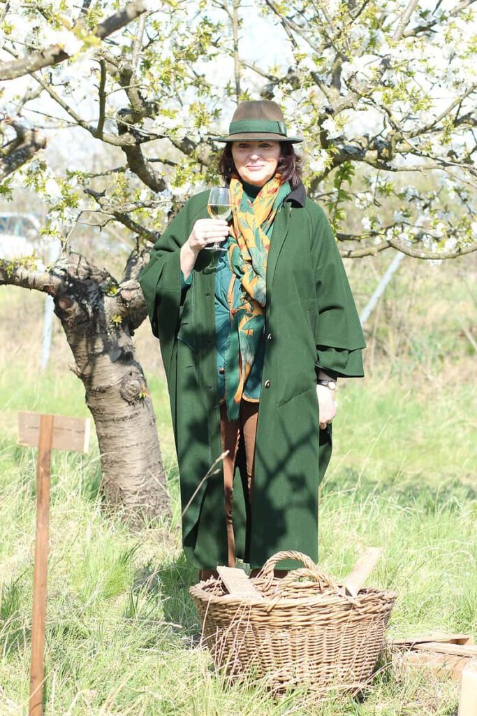 Frau im grünen Mantel steht mit einem Glas Wein im Kirschgarten